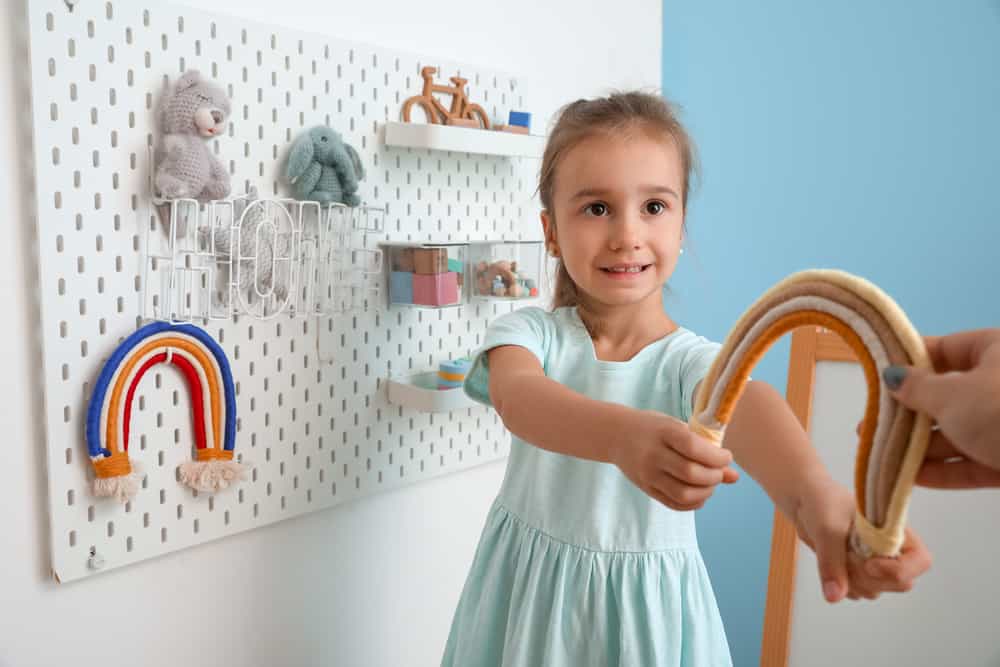 Cute little girl with toy rainbow near hanging peg board on light wall