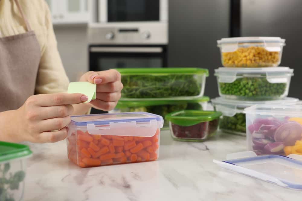 Woman sticking paper note onto container with fresh carrots at white marble table in kitchen, closeup. Food storage