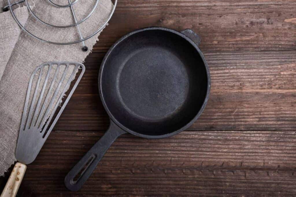 A cast iron skillet on a wooden surface alongside a metal spatula and a cloth.