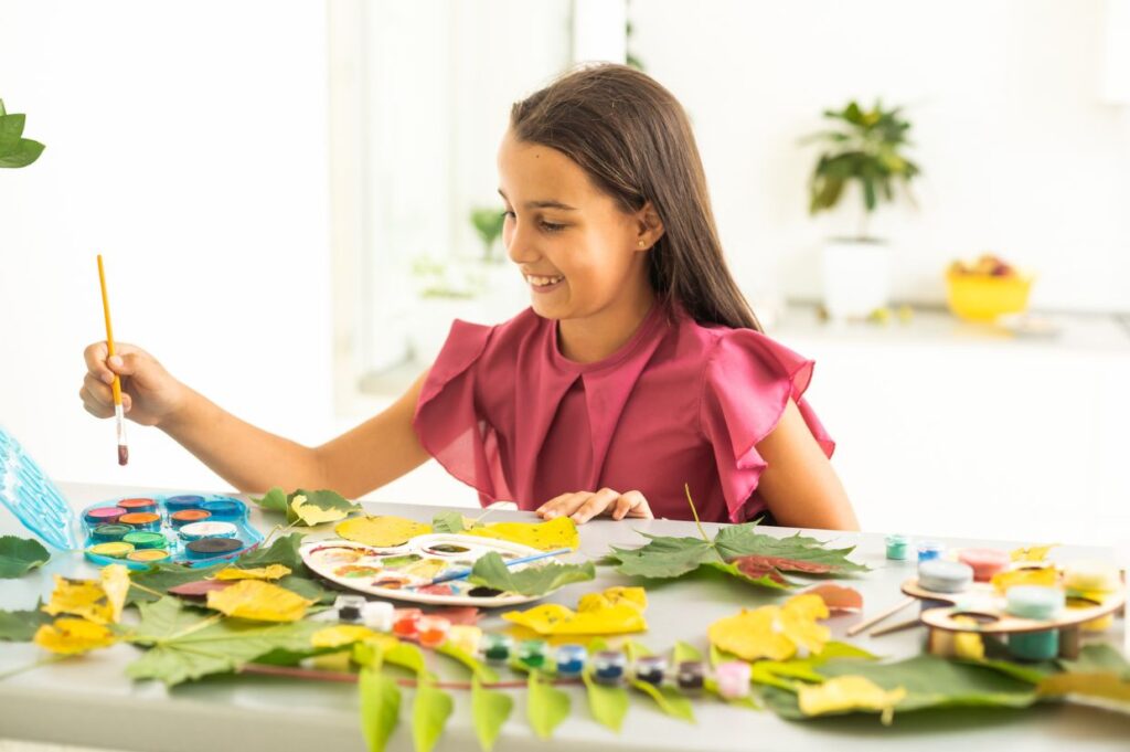 A young girl is sitting at a table, painting leaves and creating an autumn craft using natural materials like colorful leaves, paint, and brushes.