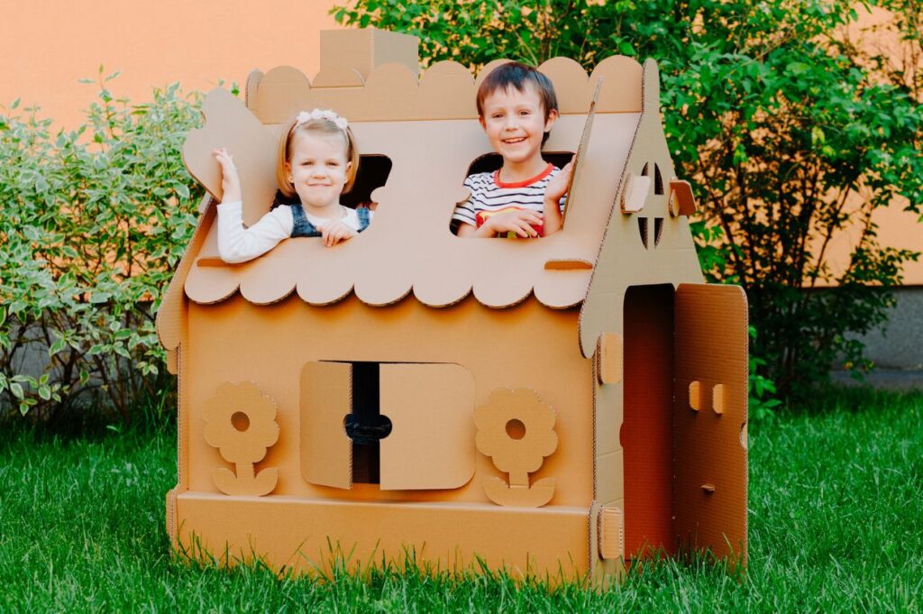 Two children are playing and smiling inside a cardboard playhouse outdoors on a green lawn. The playhouse has cut-out windows, doors, and decorative flowers.