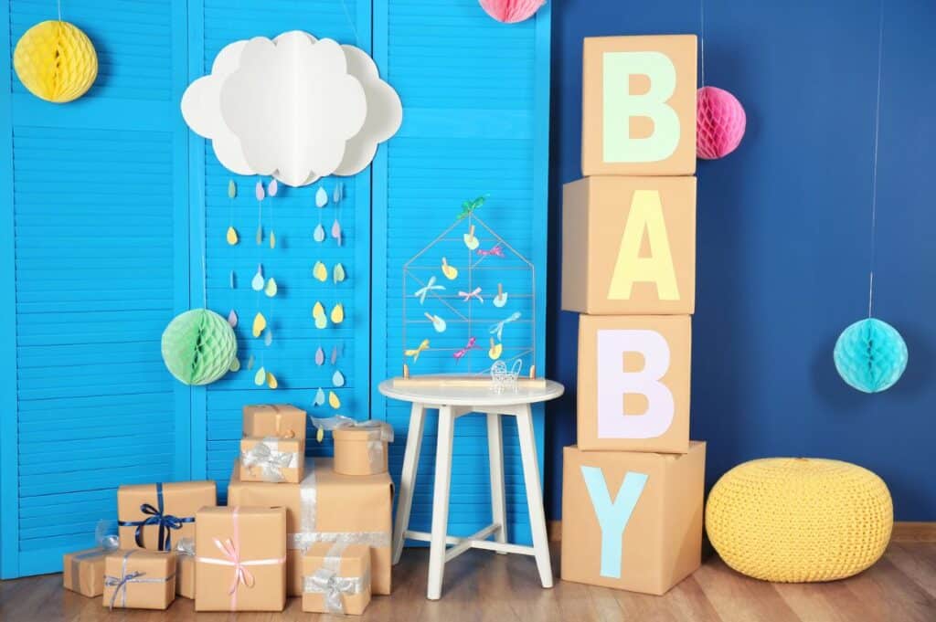 A colorful baby shower setup featuring large cardboard boxes spelling 'BABY,' a cloud and raindrop mobile, wrapped gift boxes with ribbons, and decorative paper orbs against a blue backdrop.