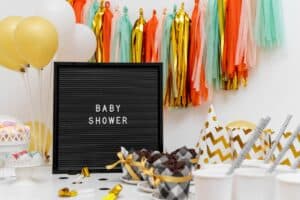 Baby shower setup with a black letter board displaying 'BABY SHOWER,' colorful tassel garlands in orange, teal, and gold, gold and white balloons, party hats, cupcakes, and decorative jars of candy.