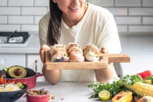 A young woman in a kitchen holding a wooden tray of fresh spring rolls in rice paper, surrounded by colorful vegetables and ingredients.