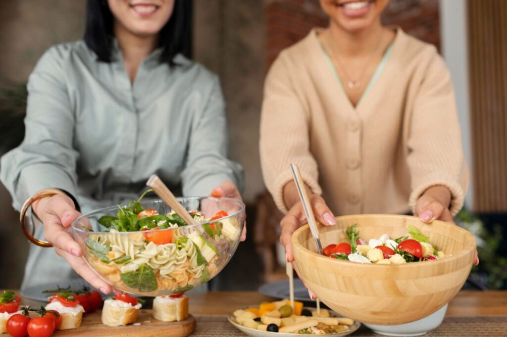 Two people are smiling and holding out bowls of fresh salad, participating in a food swap event with various appetizers on the table in front of them.