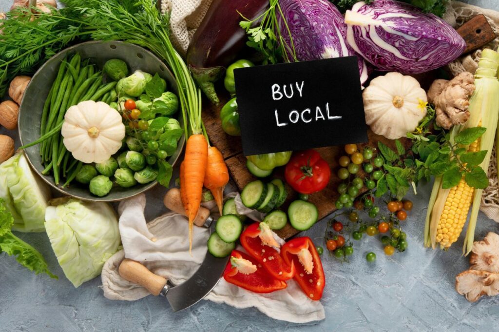 A colorful assortment of fresh local vegetables displayed on a light surface, including carrots, green beans, Brussels sprouts, cabbage, red peppers, tomatoes, cucumbers, corn, eggplant, and mini pumpkins.