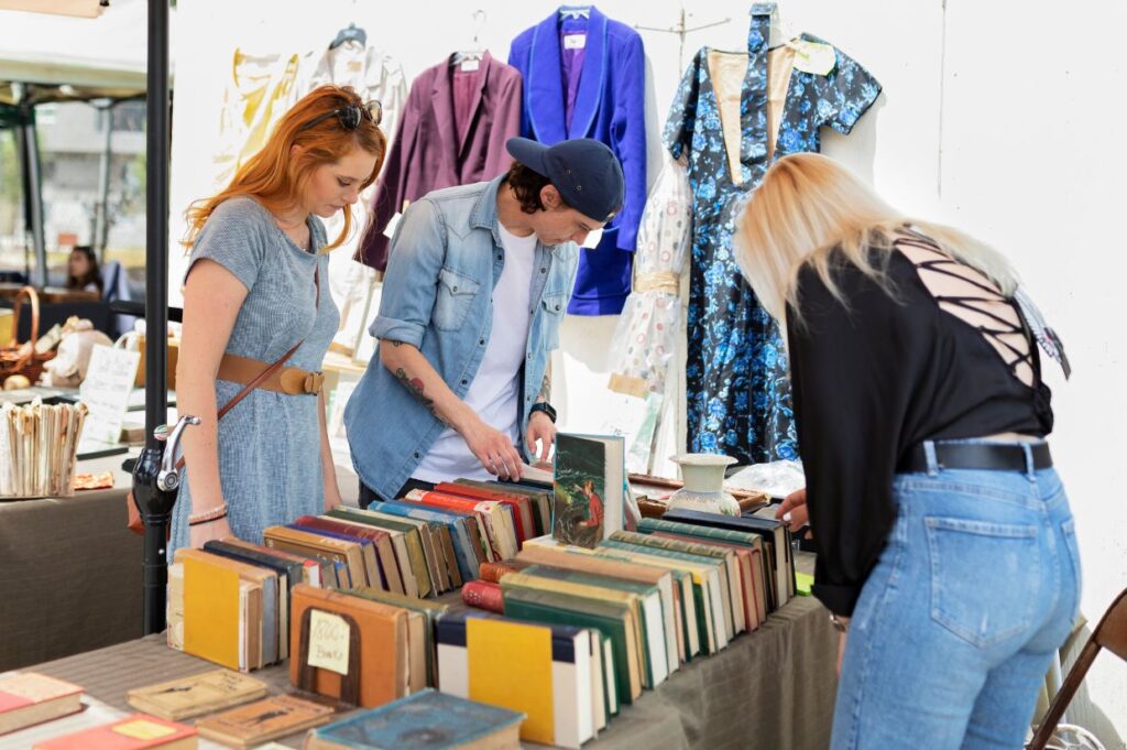 Three people are browsing a variety of old books displayed on a table at an outdoor book swap event, with vintage clothing hanging in the background.